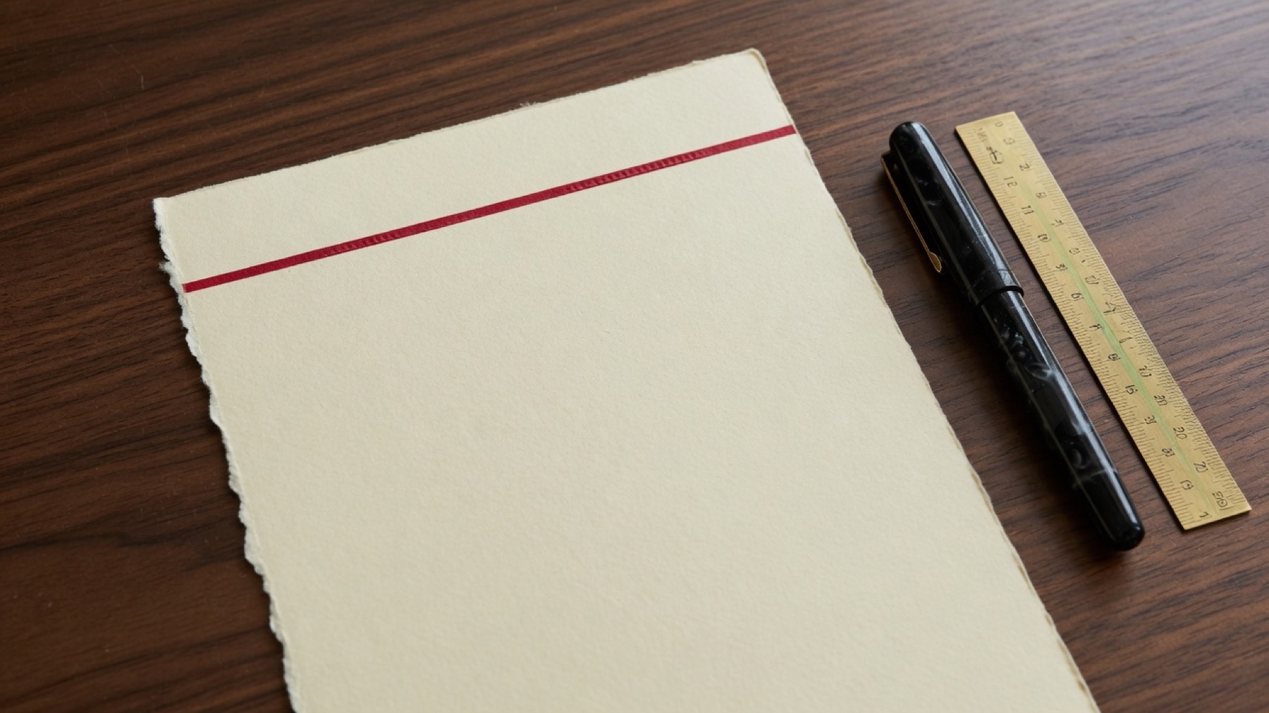 A blank cream sheet with a crimson rule on a walnut desk, next to a fountain pen and a brass ruler.